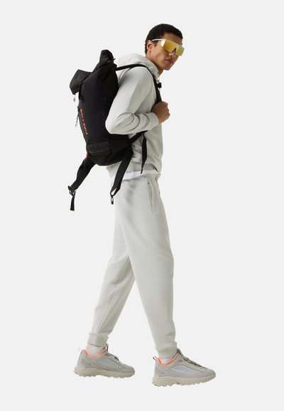 Man draagt een lichtgrijs trainingspak, beige sneakers, geelgetinte zonnebril en draagt een zwarte rugzak met het label "FIRE+ICE" over één schouder.