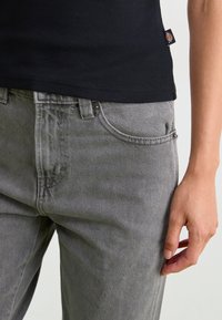Vaqueros de mezclilla gris con un corte clásico, que presentan un diseño de cinco bolsillos, cierre de botón metálico y detalle de costura. Top de tirantes negro con textura, usado por encima.
