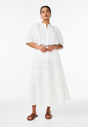 Nainen seisoo valkoisessa pitkässä hameessa ja siihen sopivassa valkoisessa pitsikoristeisessa paidassa, yhdistettynä ruskeisiin tasapohjaisiin sandaaleihin.