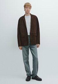 Bruine suède blazer met twee voorzakken, gedragen over een lichtgrijze trui en gecombineerd met lichtblauwe spijkerbroeken en zwarte schoenen.