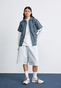 Camisola de manga curta às riscas em azul e branco, usada sobre uma camisa branca de manga comprida, combinada com calções azuis claros até ao joelho e ténis.