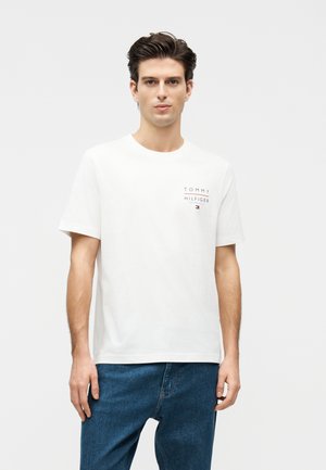 Młody mężczyzna o ciemnych włosach, ubrany w białą koszulkę Tommy Hilfiger i niebieskie jeansy, stoi na jednolitym jasnym tle.