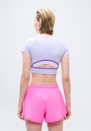 Personne aux cheveux blonds courts portant un haut court violet clair avec un dos découpé et un short rose vif, debout de dos sur fond blanc.