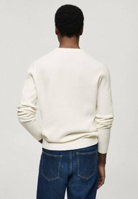 Pull en crème à côtes, avec des manches longues, un col rond et des poignets ajustés, assorti à un jean en denim bleu foncé. La texture est lisse et ajustée.