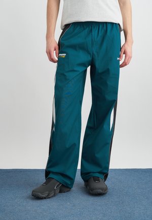 Person trägt türkise Trainingshose mit schwarzen und weißen Seitenstreifen und schwarze Slipper, steht auf blauem Teppichboden.