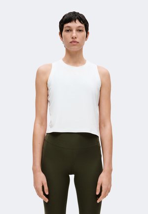 Personne portant un top blanc sans manches et court ainsi que des leggings taille haute vert olive, debout sur un fond gris clair.