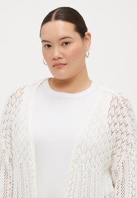 White knitted cardigan with a lace pattern, open front design, and long sleeves, worn over a white ribbed shirt.