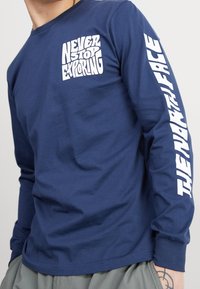 Marinblå långärmad skjorta i bomull med vit text: "SLUTA ALDRIG UTFORSKA" framtill och "THE NORTH FACE" längs ärmen.