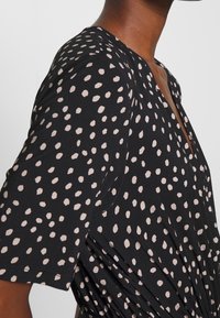 Robe noire avec un décolleté en V et des manches courtes, ornée d'un motif de pois crème. Les détails froncés ajoutent de la texture au tissu.