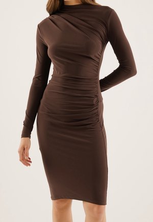 Femme portant une robe moulante marron foncé à manches longues, froncée, longueur genou avec un col montant.