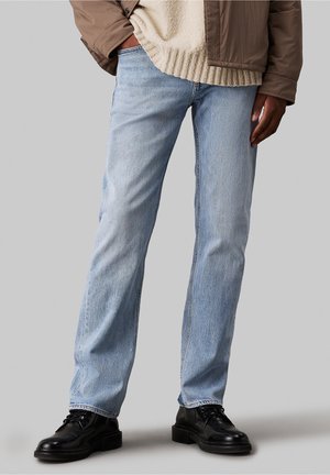 Personne portant un jean bleu clair droit, des chaussures noires à lacets, un pull beige texturé et une veste marron avec une main dans la poche.
