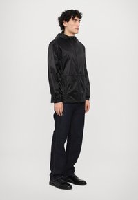 Veste noire, légère, à capuche avec fermeture éclair sur le devant, manches froncées et ourlet élastique, associée à un jean foncé et des chaussures noires.