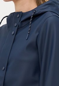 Voděodolná námořnicky modrá bunda s přiléhající kapucí, zapínáním na druky a nastavitelným stahovacím šňůrkou. Hladká textura a bezešvý design.