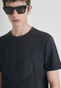 Mladić nosi crne sunčane naočale i crnu majicu sa suptilnim reljefnim dizajnom tigrove glave na prednjoj strani.