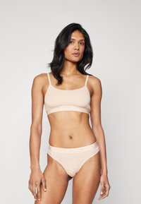 Bež bralette in tanga komplet z tanjšimi naramnicami, ki vključuje kontrastno logotipno elastično pasu. Gladka tekstura in prilegajoča se oblika.