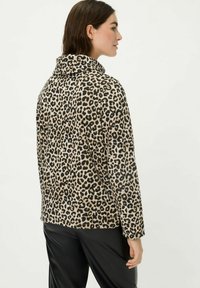 Frau in einer Jacke mit Leopardenmuster und schwarzen Hosen, von hinten gezeigt, vor einem schlichten hellen Hintergrund stehend.