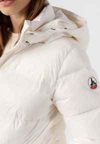 Witte donsjas met capuchon, met een gladde textuur en doorgestikte afwerking. Voorzien van een logopatch op de linker mouw en ritsdetails.