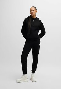 Zwarte hoodie en joggers van zacht materiaal, met een voorzak en logodetail. Gepaard met witte sneakers en geribde sokken.