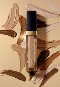 Estée Lauder Futurist concealer i en genomskinlig tube med ett svart lock, omgiven av färgprover av olika hudtoner i foundation på en guldig bakgrund.