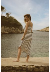 Vestito midi a righe bianche e nere con spalline sottili, spacco laterale, orlo svasato e tessuto leggero, indossato a piedi nudi da una donna vicino all'acqua.