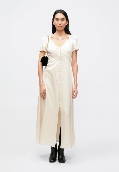 Κρεμ φόρεμα από σατέν με μπούστο από δαντέλα, κοντά φουσκωτά μανίκια και κούμπωμα με κουμπιά μπροστά. Διαθέτει σκίσιμο μέχρι τον μηρό και φοριέται με μαύρα μποτάκια αστραγάλου.