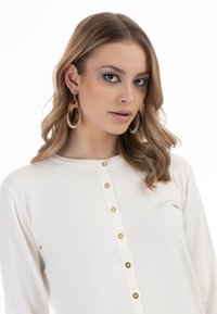 Bílé pletené tričko s dlouhými rukávy a kulatým výstřihem, zdobené pěti zlatými knoflíky vpředu. Modelka má na sobě velké průhledné kruhové náušnice.