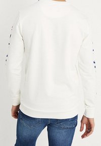 Sudadera blanca de manga larga, con puños y dobladillo acanalados. Presenta letras en las mangas en azul marino y rojo. Tejido de mezcla de algodón, ajuste casual.