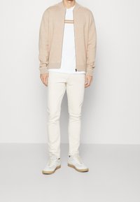 Giacca bomber a maglia beige sopra una camicia bianca con due righe orizzontali. Jeans chiari e sneakers bianche con suola beige.