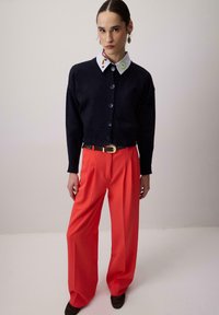 Cardigan court bleu marine avec de gros boutons, associé à un pantalon large orange vif et une ceinture noire avec une boucle dorée. Col blanc brodé.
