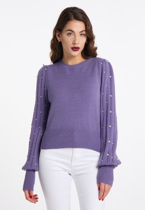 Donna dai capelli scuri che indossa un maglione viola con dettagli di perle sulle maniche a sbuffo e pantaloni bianchi su sfondo bianco.