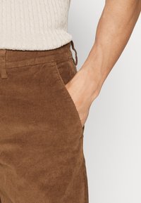 Bruna kordflandy med en ribbad struktur, med sidofickor och en avslappnad passform. Synlig dragsko i midjan för justering.