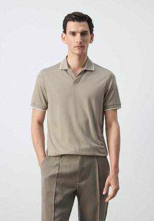 Un homme se tient face à l’avant portant un polo beige à manches courtes et un pantalon assorti, la main droite dans la poche sur un fond uni.