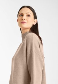 Beige gebreide trui met een hoge geribbelde kraag, verlaagde schouders en een losse pasvorm. Zachte textuur, minimalistisch ontwerp en neutrale kleur.