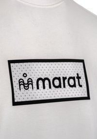 Witte T-shirt met daarop een rechthoekig, doorzichtig label met zwarte rand waarop het woord "marat" staat en een golvend logo, geplaatst tegen een stippenachtergrond.