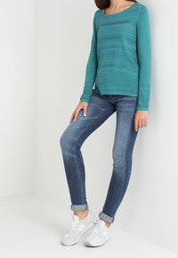 Femme portant un haut à manches longues turquoise, un jean skinny bleu retroussé aux chevilles et des baskets blanches, debout contre un mur blanc.