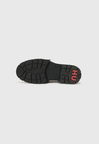 Suola della scarpa, in gomma nera con battistrada profondo, presenta un prominente logo rosso "HU" sul tallone. Superficie testurizzata per aderenza e durata.