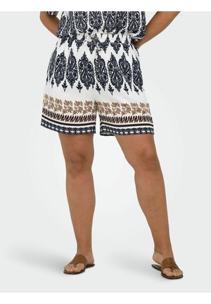 Persona que lleva pantalones cortos blancos con patrones intrincados en negro y marrón, una blusa a juego y sandalias marrones con los dedos al descubierto, de pie con las manos en las caderas.