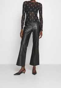 Schwarzes, transparentes Langarmoberteil mit wiederholtem Logo-Print, kombiniert mit hochtaillierten, schwarzen Leder-Hosen mit Schlag und schwarzen Slingback-Absätzen.