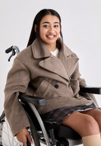 Młoda kobieta z długimi ciemnymi włosami uśmiechająca się, siedząca na wózku inwalidzkim, ubrana w brązowy płaszcz i kraciastą spódnicę na jednolitym tle.