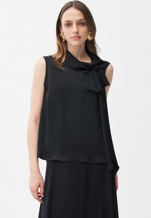 Femme portant un chemisier noir sans manches avec un nœud noué au col et de grandes boucles d'oreilles rondes dorées, debout devant un fond uni.