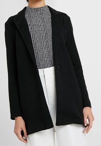 Černý blazer s texturovanou tkaninou, přiléhavý střih, zapínání na jedno tlačítko a dvěma předními kapsami, doplněný vzorovaným topem a bílými kalhotami.