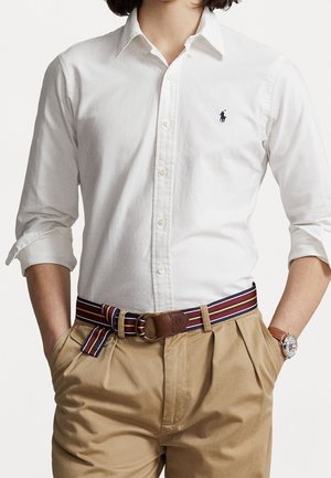 Homme portant une chemise blanche à manches longues avec un logo foncé, une ceinture en tissu rayée avec boucle en cuir, un pantalon beige et une montre-bracelet, mains dans les poches.
