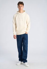 Jonge man met een crèmekleurige hoodie, donkerblauwe spijkerbroek en witte sneakers staat met één hand in zijn zak tegen een effen lichte achtergrond.