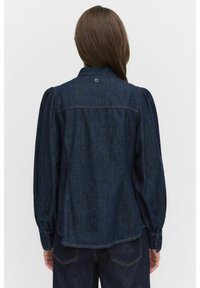Jeansskjorta i mörkblått med puffärmar, krage och subtila sömmar. Har en cirkulär detalj på baksidan.