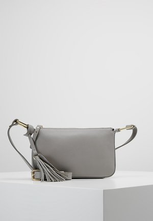 Sac bandoulière en cuir gris avec sangle ajustable, ferrures dorées, fermeture éclair et pompon décoratif placé sur une surface blanche.
