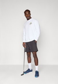 Camicia bianca a maniche lunghe con logo, pantaloni corti neri, scarpe sportive blu e calze bianche; in mano tiene una mazza da golf; in piedi su uno sfondo neutro.