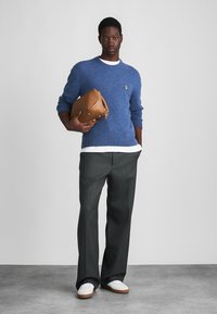 Μπλε μάλλινο πουλόβερ με λευκό υποστρώμα, γκρι υφαντές παντελόνες, λευκά αθλητικά παπούτσια και καφέ δερμάτινη τσάντα κρατημένη στο ένα χέρι.