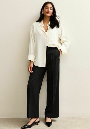 Blusa de seda color crema con un bolsillo, combinada con pantalones de pierna ancha negros y tacones style slingback negros. Textura suave y corte relajado.