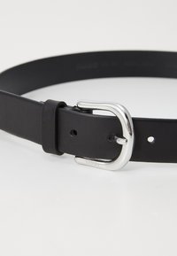 Svart lærbelte med en glatt tekstur, med en sølvfarget metallspenne og flere hull for justering. Pr präget logo detalj på spennen.