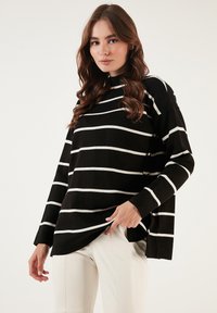 Pulover cu dungi alb-negru, cu mâneci lungi, guler înalt și croială largă, realizat dintr-un material moale cu textură vizibilă și tiv rotunjit.
